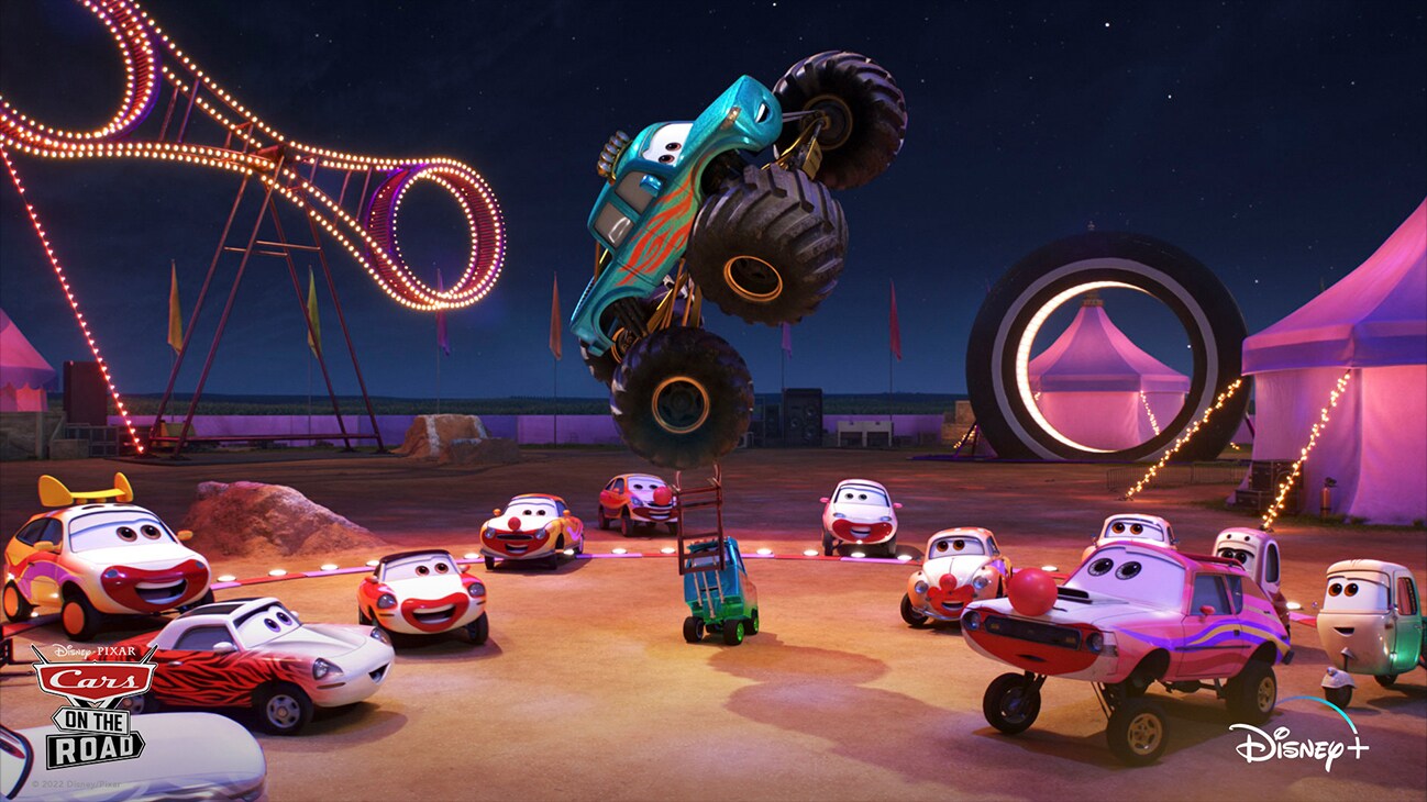 A car with monster truck sized wheels is balanced in the air by a forklift at a fair surrounded by several hot rod cars from the Disney+ Original series "Disney•Pixar Cars on the Road".