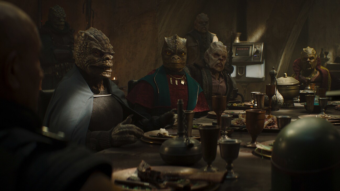(L): Boba Fett (Temura Morrison) sitting at a table with several alien characters in Lucasfilm's THE BOOK OF BOBA FETT, exclusively on Disney+. © 2021 Lucasfilm Ltd. & ™. All Rights Reserved.