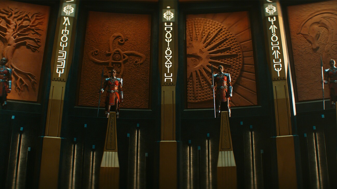 The Dora Milaje stand in a circular chamber atop pedestals, with intricate wall carvings behind them. From the film, Marvel Studios' Black Panther: Wakanda Forever.