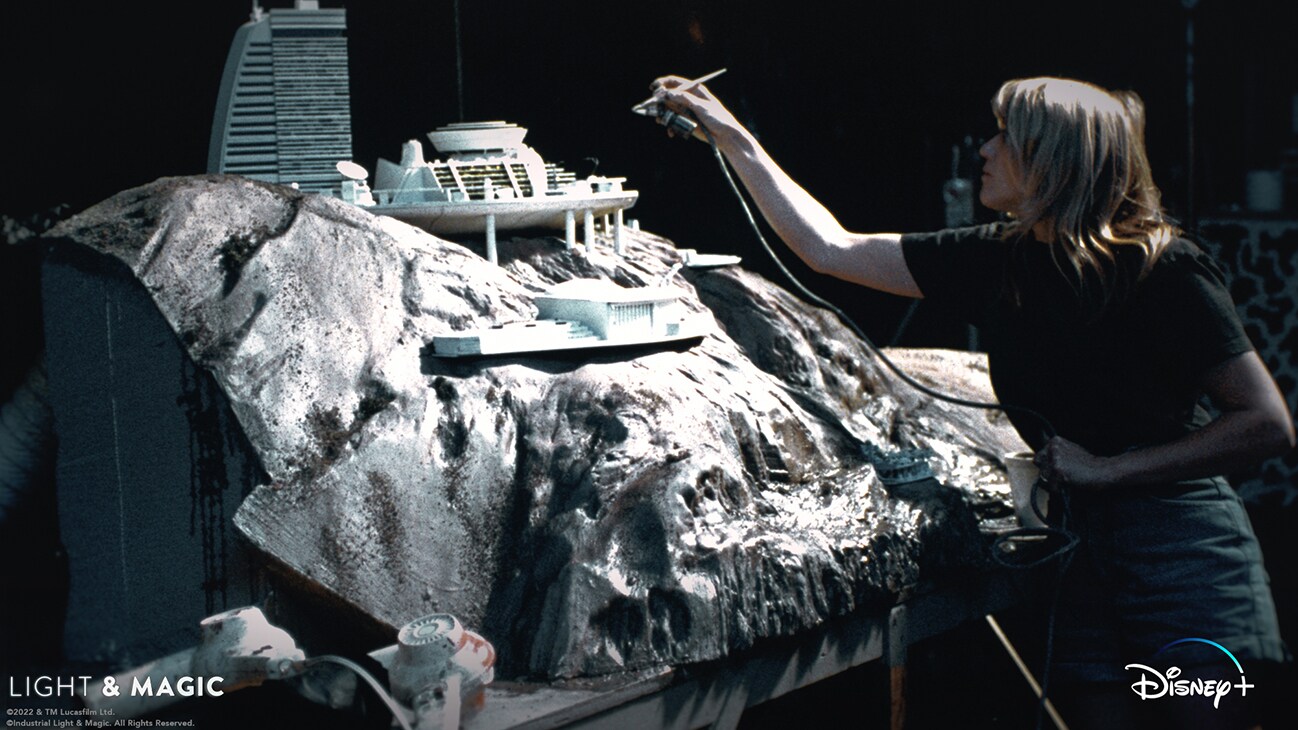 ILM artist using an airbrush on a large model of a house built into a hill from the Disney+ Original series, "Light & Magic". | © & ™ Lucasfilm Ltd. ©Industrial Light & Magic. All Rights Reserved.