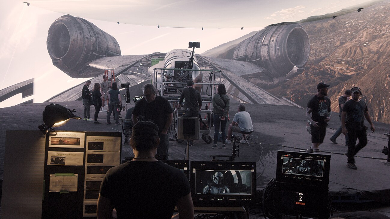 Image of a person at a computer in front of crew filming a scene from "The Mandalorian", with the Razorcrest starship on a large screen in front of them. From the Disney+ Original series "Light & Magic". | © & ™ Lucasfilm Ltd. ©Industrial Light & Magic. All Rights Reserved.