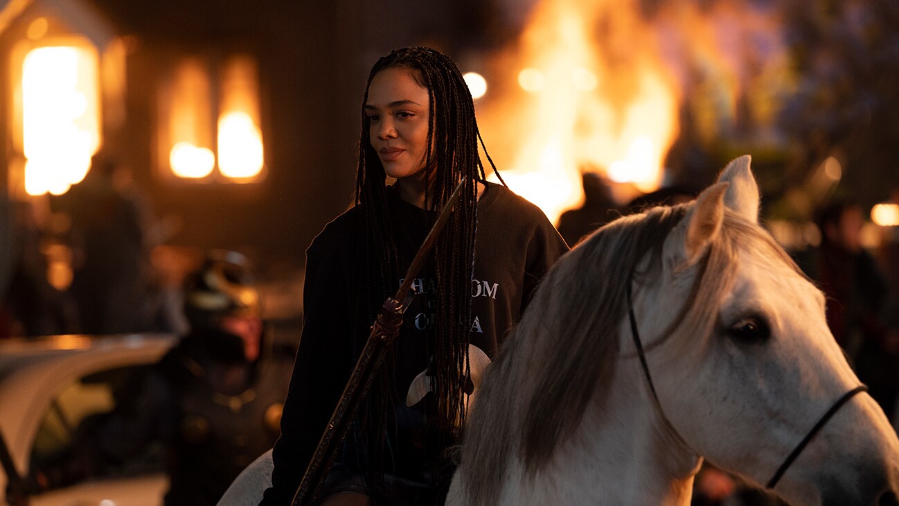 Valkyrie (actor Tessa Thompson) rides a white horse as buildings can be see on fire behind her in Marvel Studios' Thor: Love and Thunder.