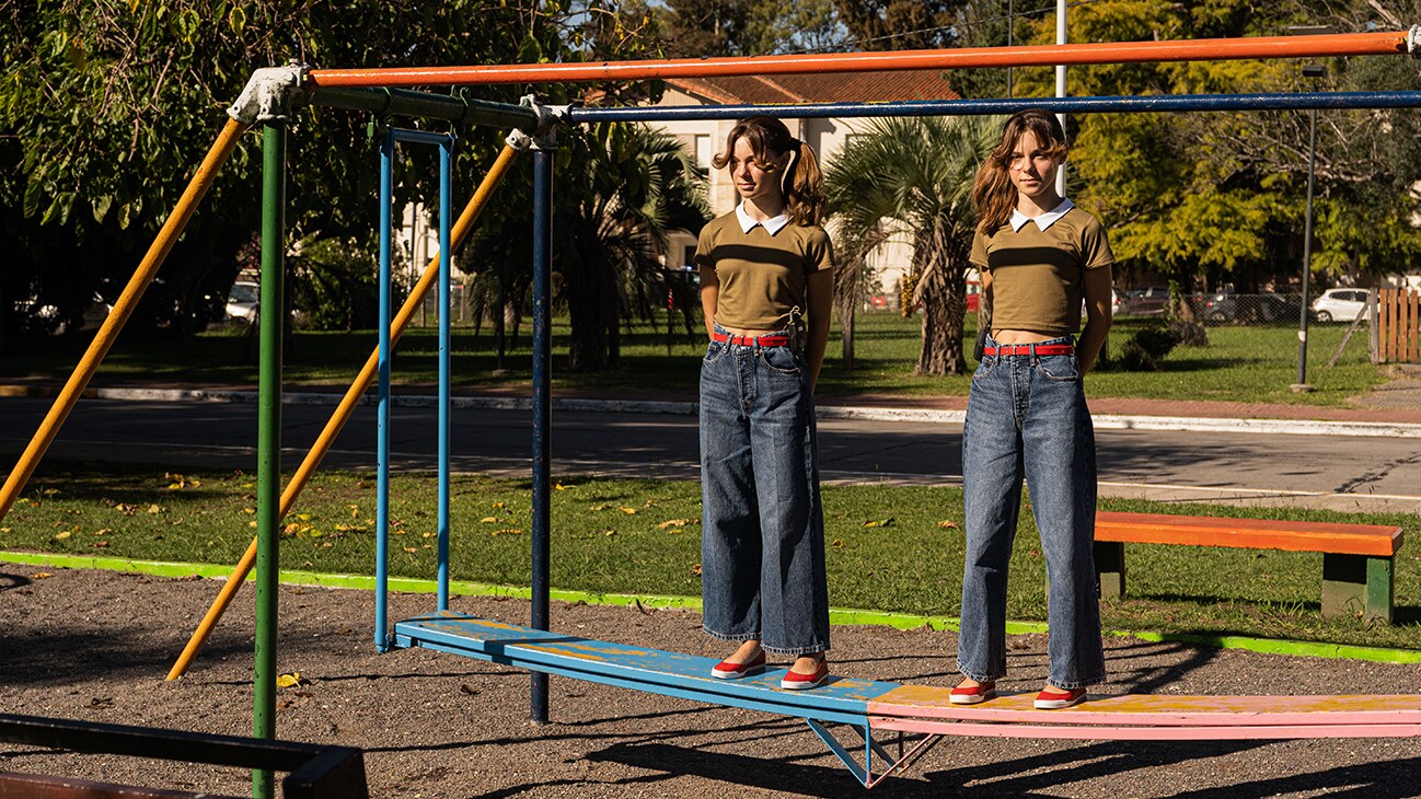 Twin girls in twin clothing stand eerily in a playground, from the Disney+ Original series, Tierra Incognita.