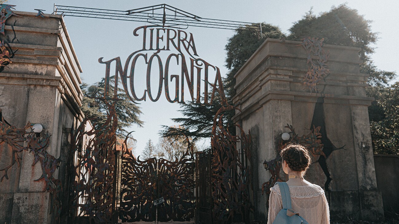 A character from the Disney+ Original series, Tierra Incognita, stands before a large gated entry to the Tierra Incognita horror theme park.