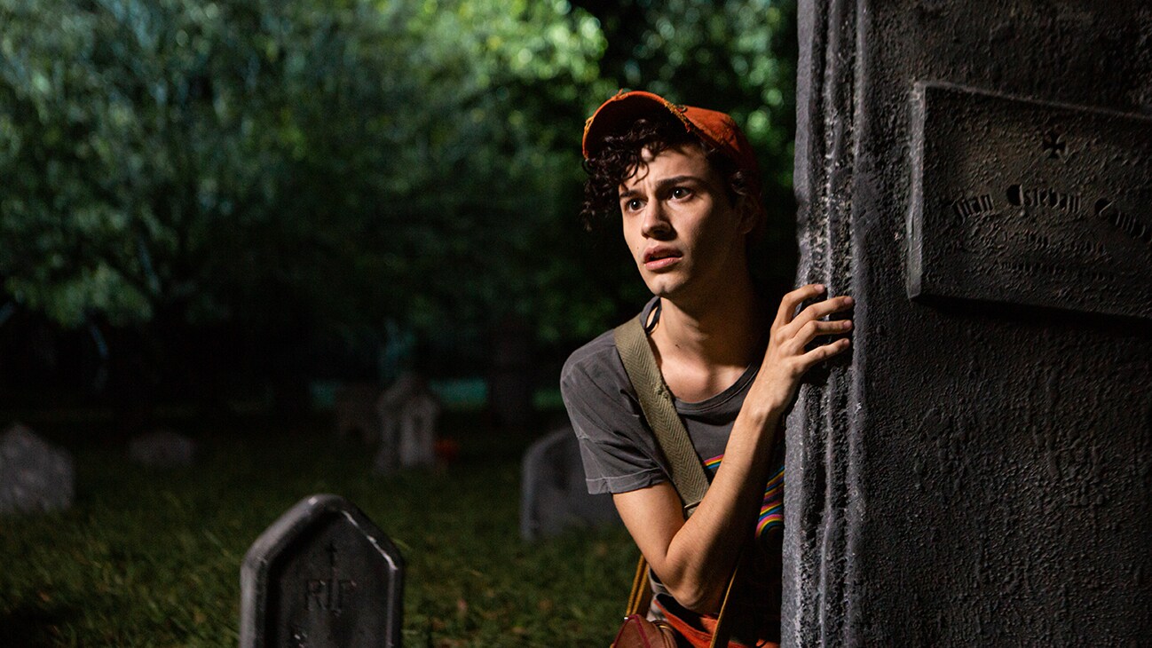 A character from the Disney+ Original series, Tierra Incognita, hides behind a tombstone in a graveyard.