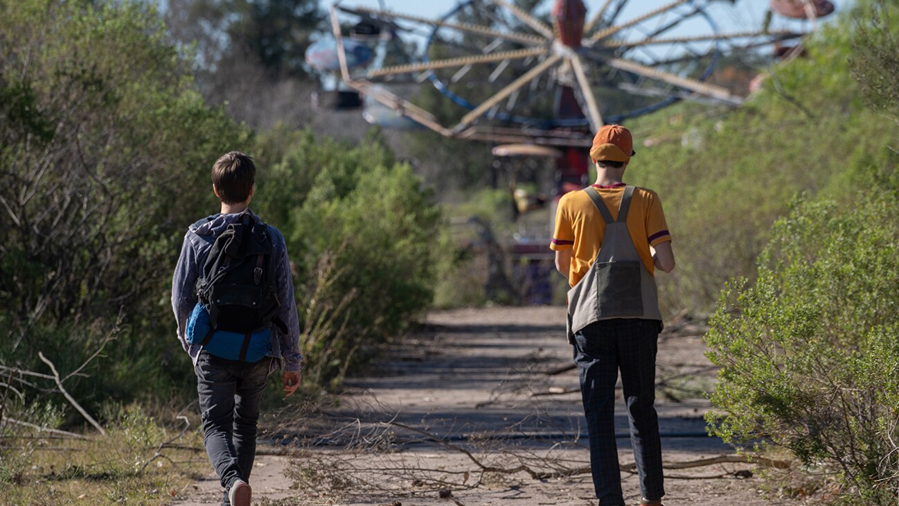 Characters from the Disney+ Original series, Tierra Incognita, walk toward an abandoned theme park attraction.