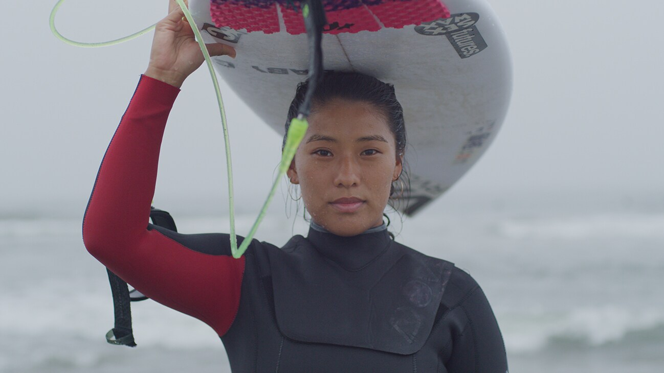 Mahina Maeda carries a surfboard above her head.
