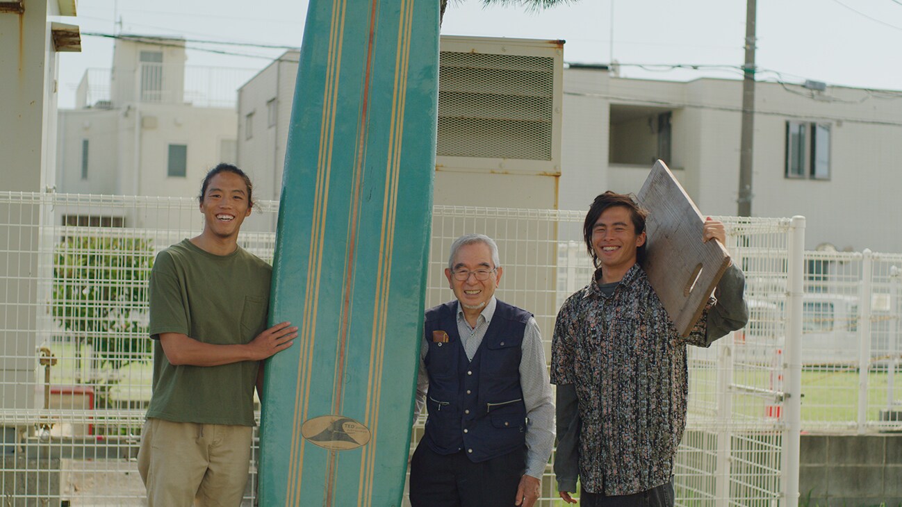 Yuma Takanuki takes a group picture with 2 others. Yuma is holding a surfboard.