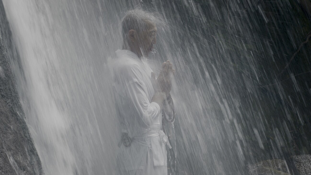 A monk stands under a waterfall. (National Geographic for Disney+)