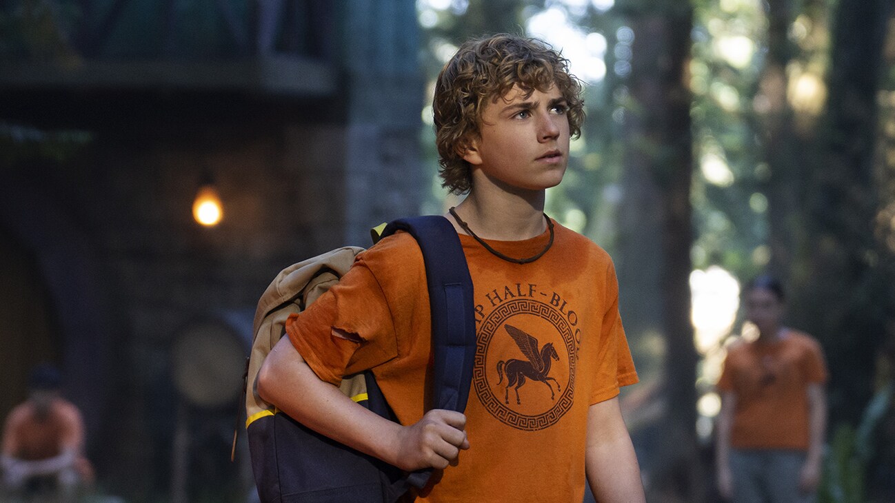 Percy Jackson (WALKER SCOBELL) wearing an orange Camp Half-Blood shirt with a backpack on his shoulder in PERCY JACKSON & THE OLYMPIANS - "Episode 102” (Disney/David Bukach).