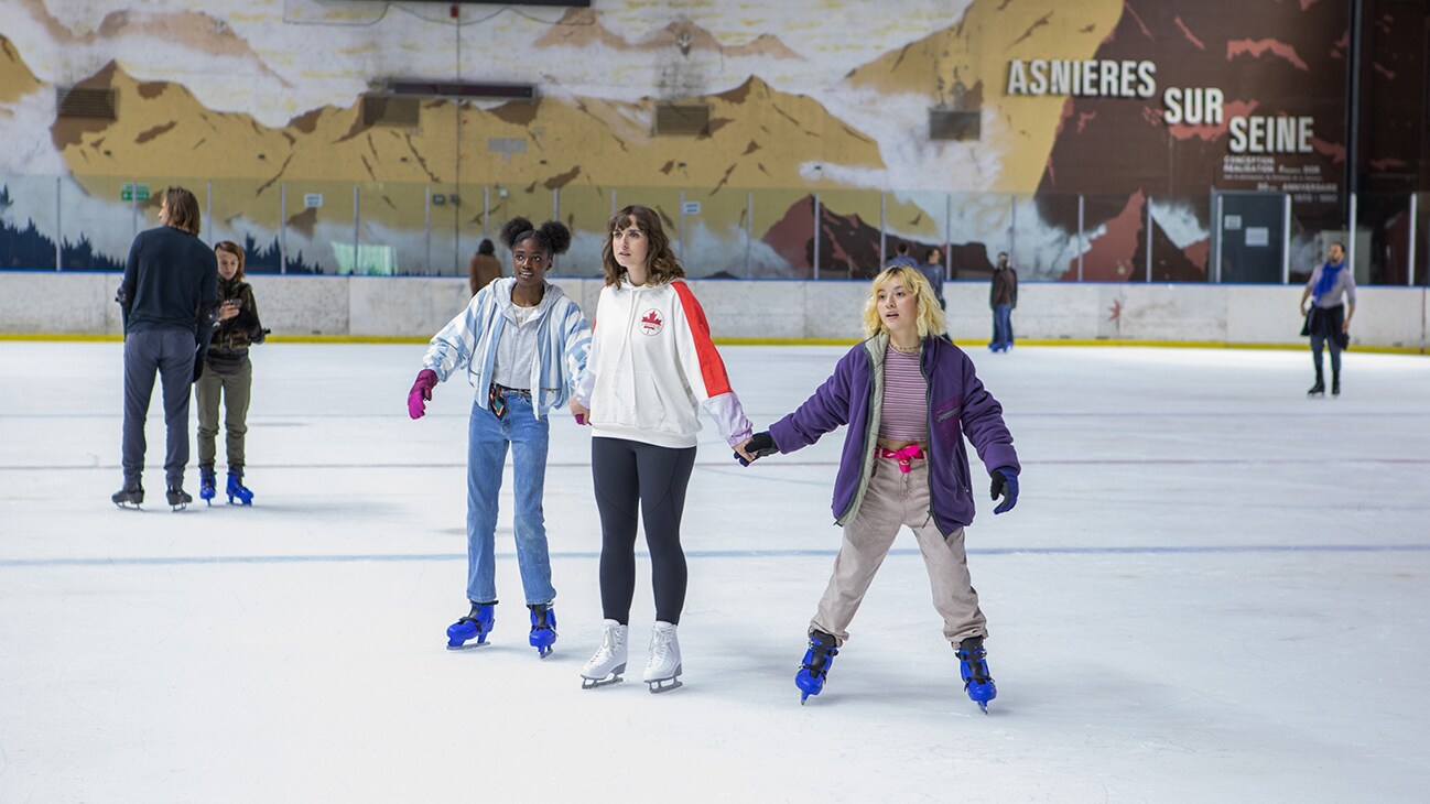 Three girls skating at an indoor ice skating rink.