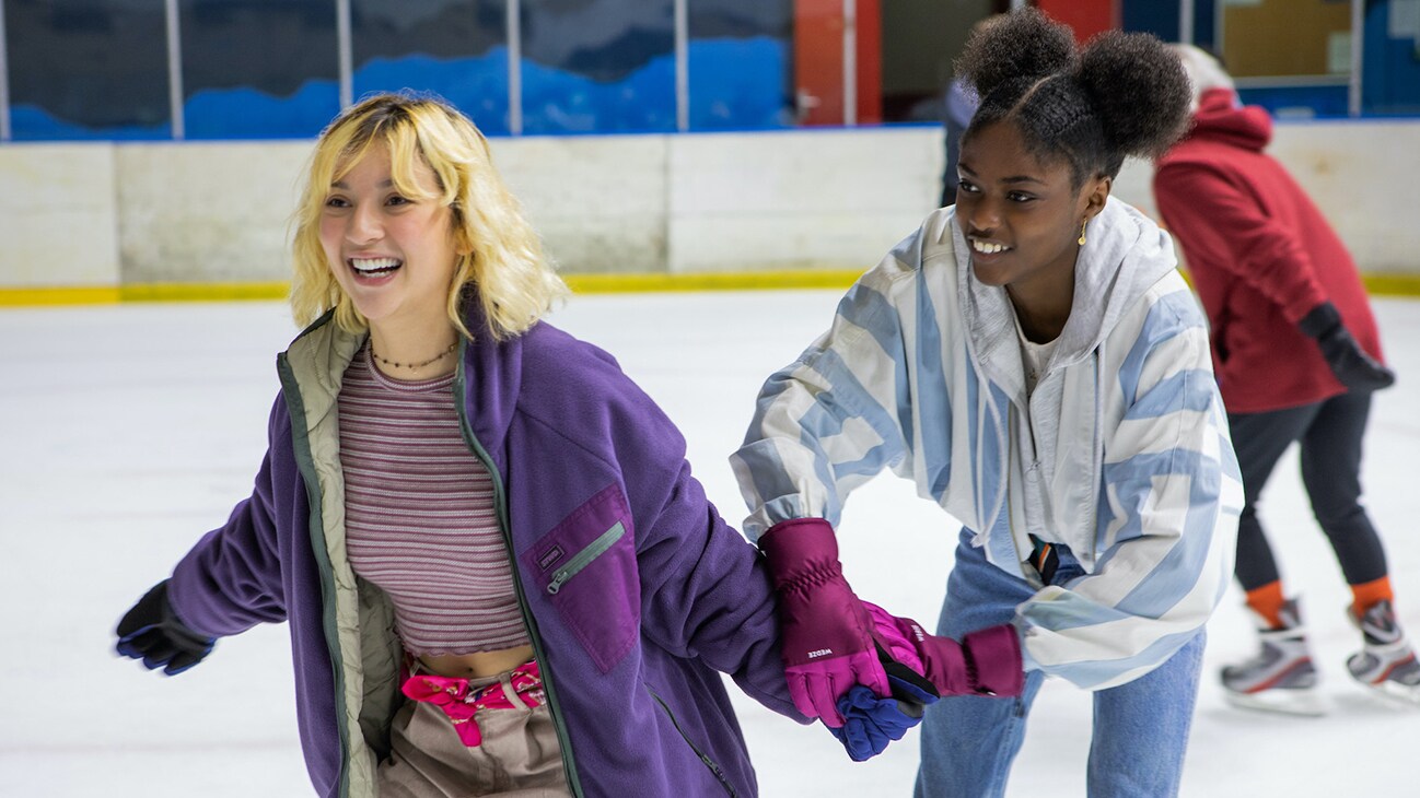 Two girls hold each other's hand as they ice skate.