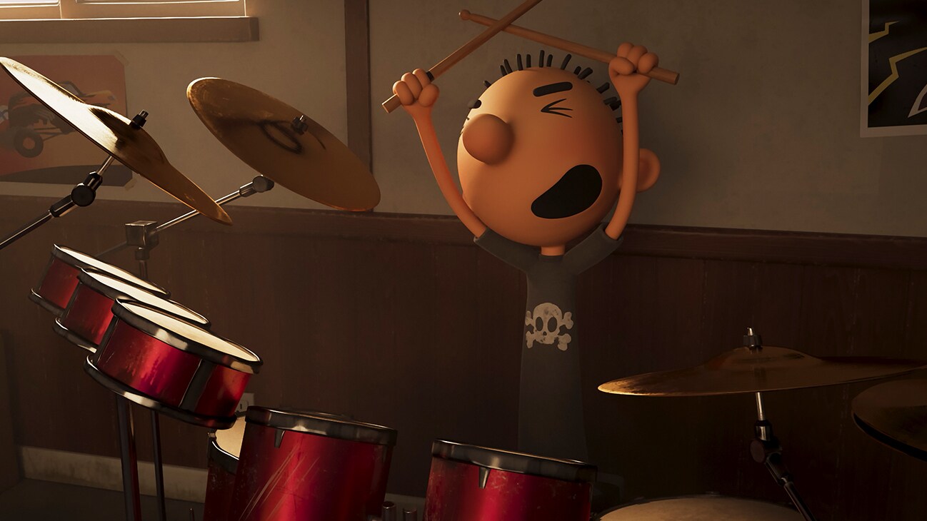 Rodrick is sitting in front of a drum set, arms stretched above him holding drumsticks with a look of joy on his face.