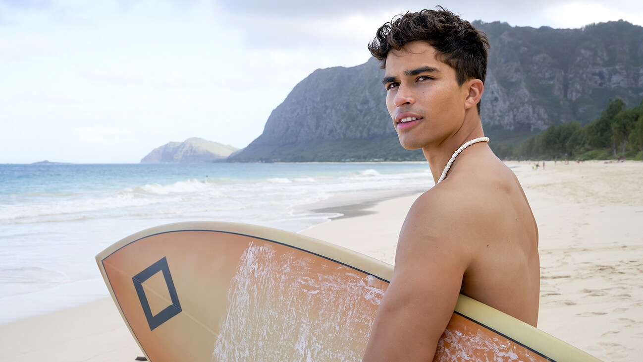 Alex Aiono as “Walter Taumata” with a surfboard on the beach from the Disney+ Original series Doogie Kamealoha, M.D.