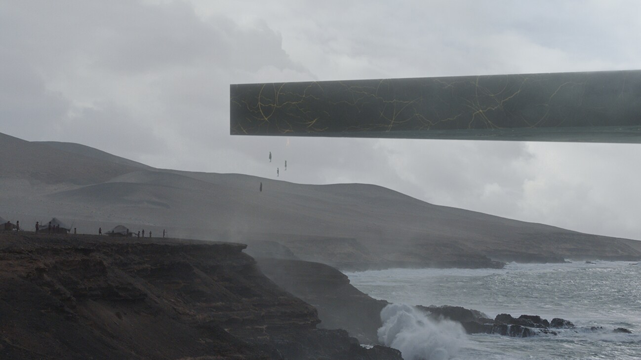 Image of a large and flat object hovering above the ocean by cliffs with several human-like objects just below it from the Marvel Studios movie Eternals.