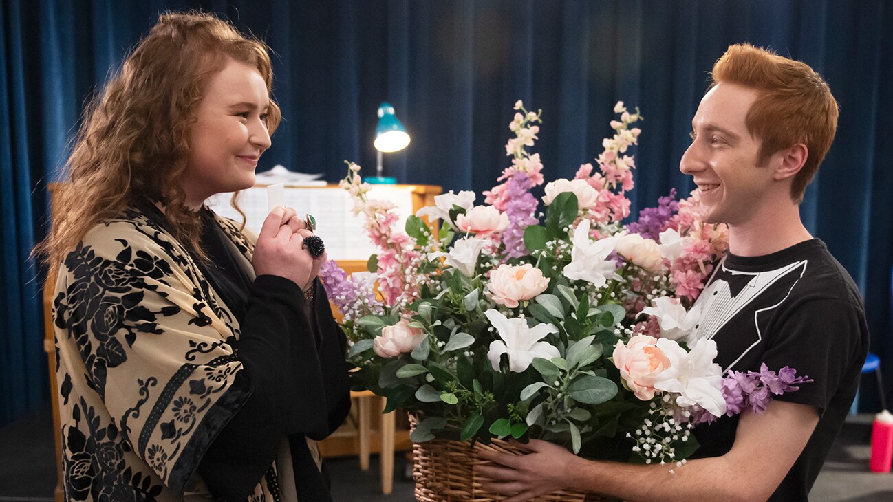 Ashlyn (actor Julia Lester) smiling staring at Big Red (actor Larry Saperstein) who is holding flowers from the Disney+ Original series "High School Musical: The Musical: The Series".