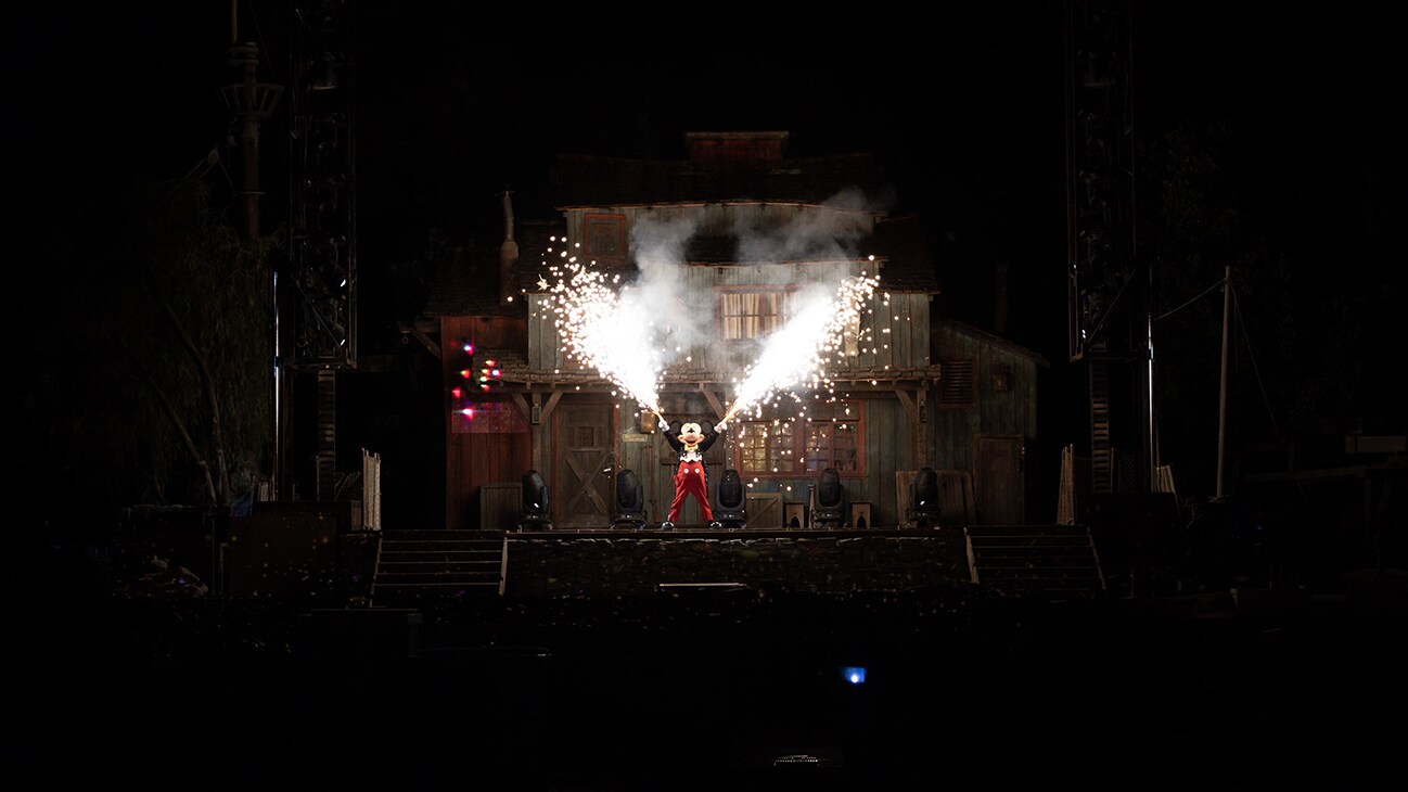 Image of Mickey Mouse on a stage with his arms up and shooting sparks from each hand.