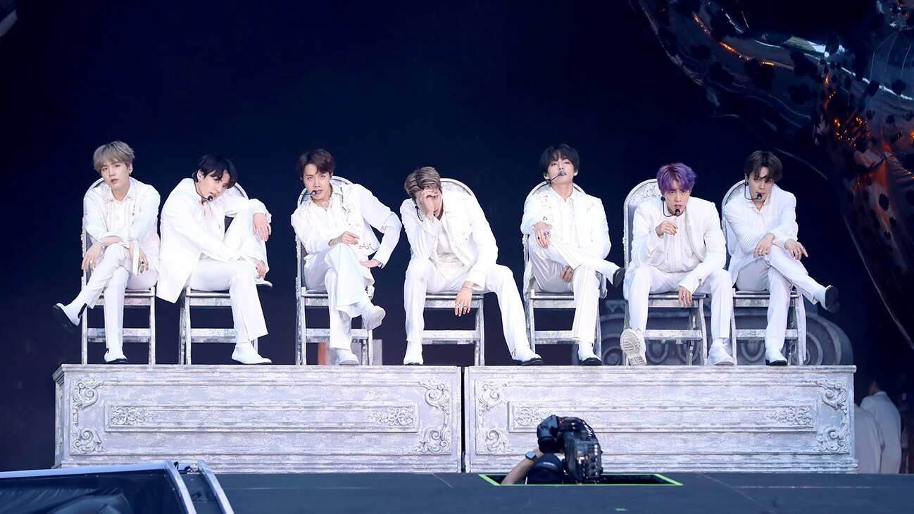 RM, Jin, SUGA, j-hope, Jimin, V and Jung Kook sitting in chairs on stage from the Disney+ docuseries, "BTS Monuments: Beyond The Star."