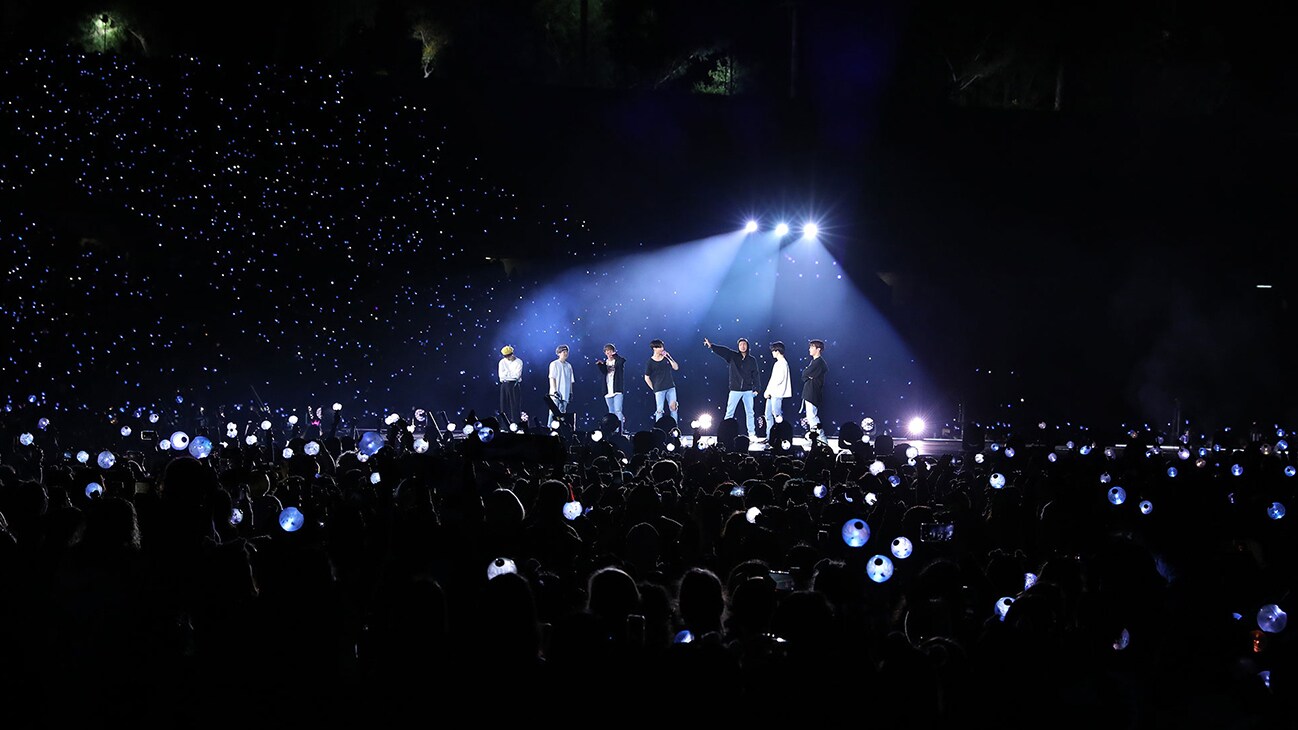 An image of a dark concert venue with a spotlight on the band from the Disney+ docuseries, "BTS Monuments: Beyond The Star."