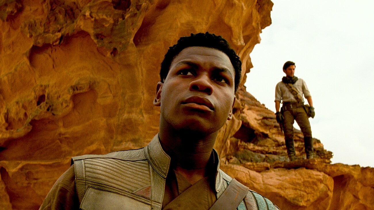 John Boyega as Finn with Oscar Isaac as Poe Dameron standing in the background, standing on a rock formation in "Star Wars: The Rise of Skywalker" 