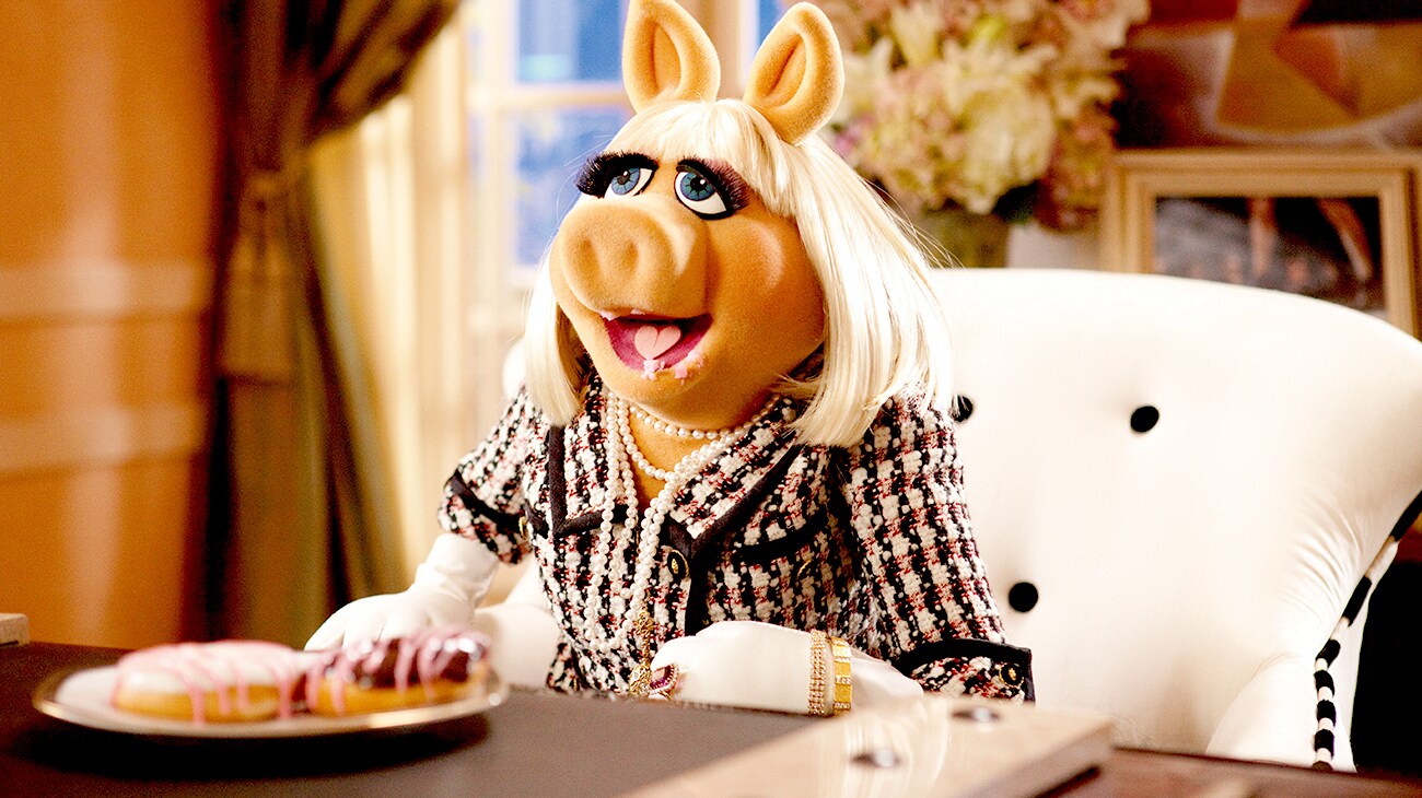 Mis Piggy voiced by Eric Jacobson sitting in front of a plate of doughnuts