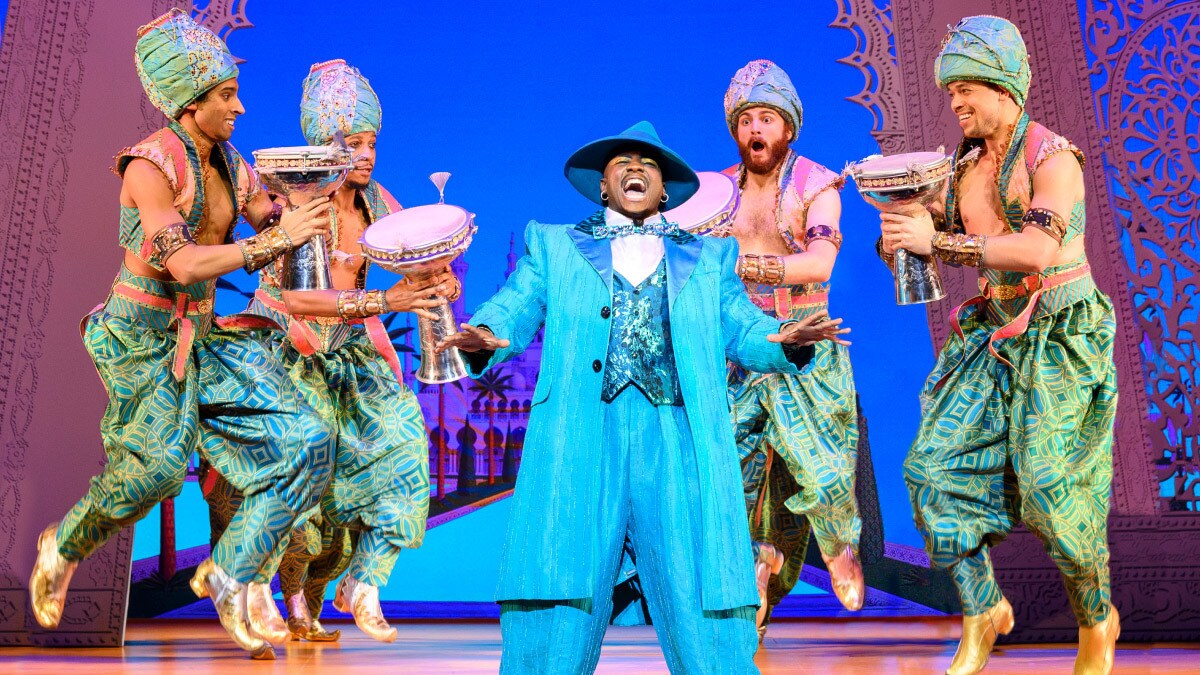 Genie in a blue suit singing among the Aladdin ensemble