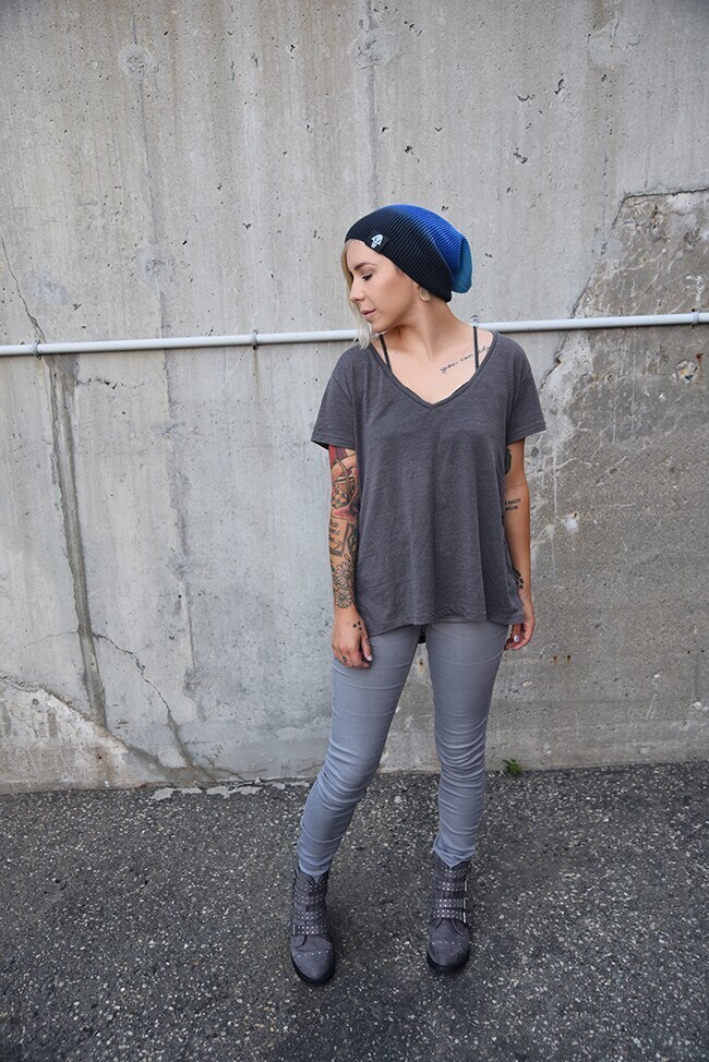 A blonde woman wearing a blue ombre-style beanie and an all-grey outfit stands against a concrete wall