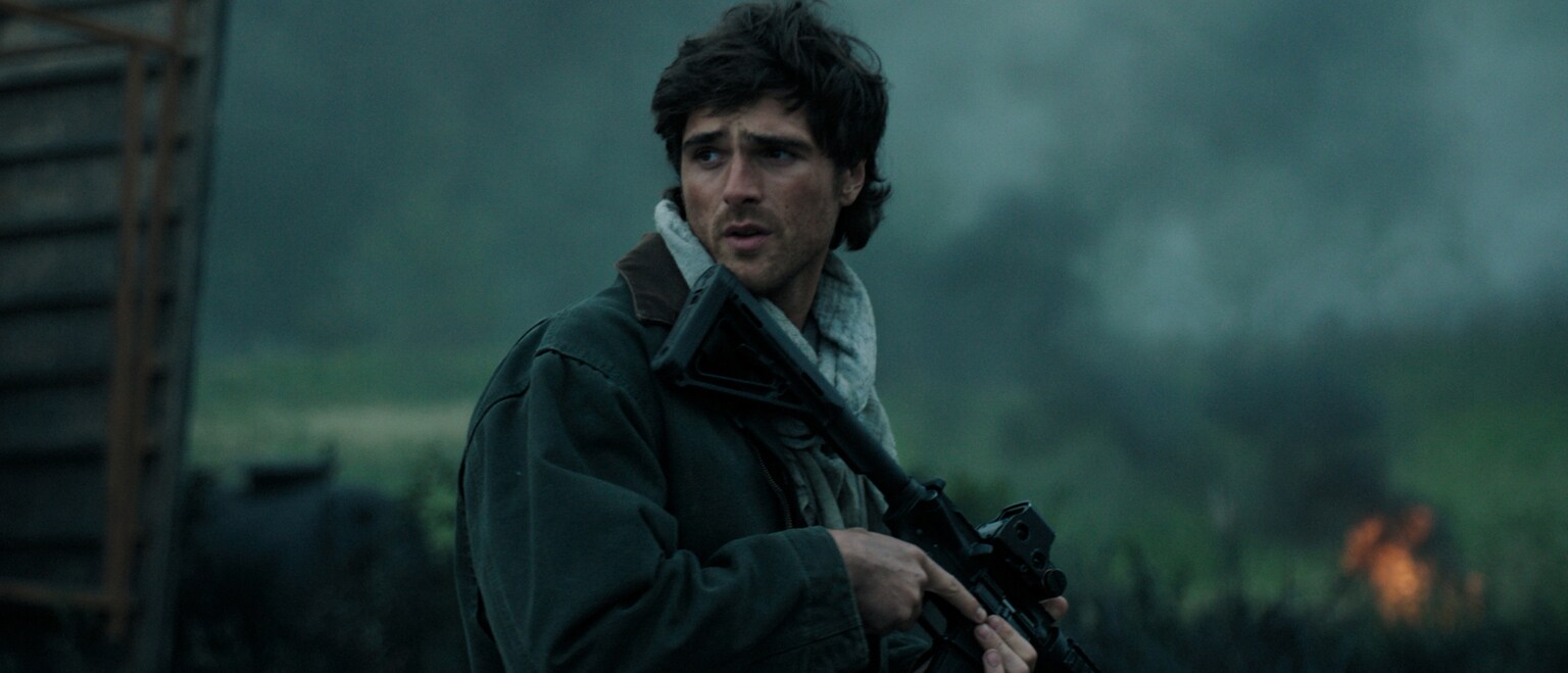 Image of actor Jacob Elordi turning to the side while holding a rifle from the 20th Century Studios movie, "The Dog Stars."