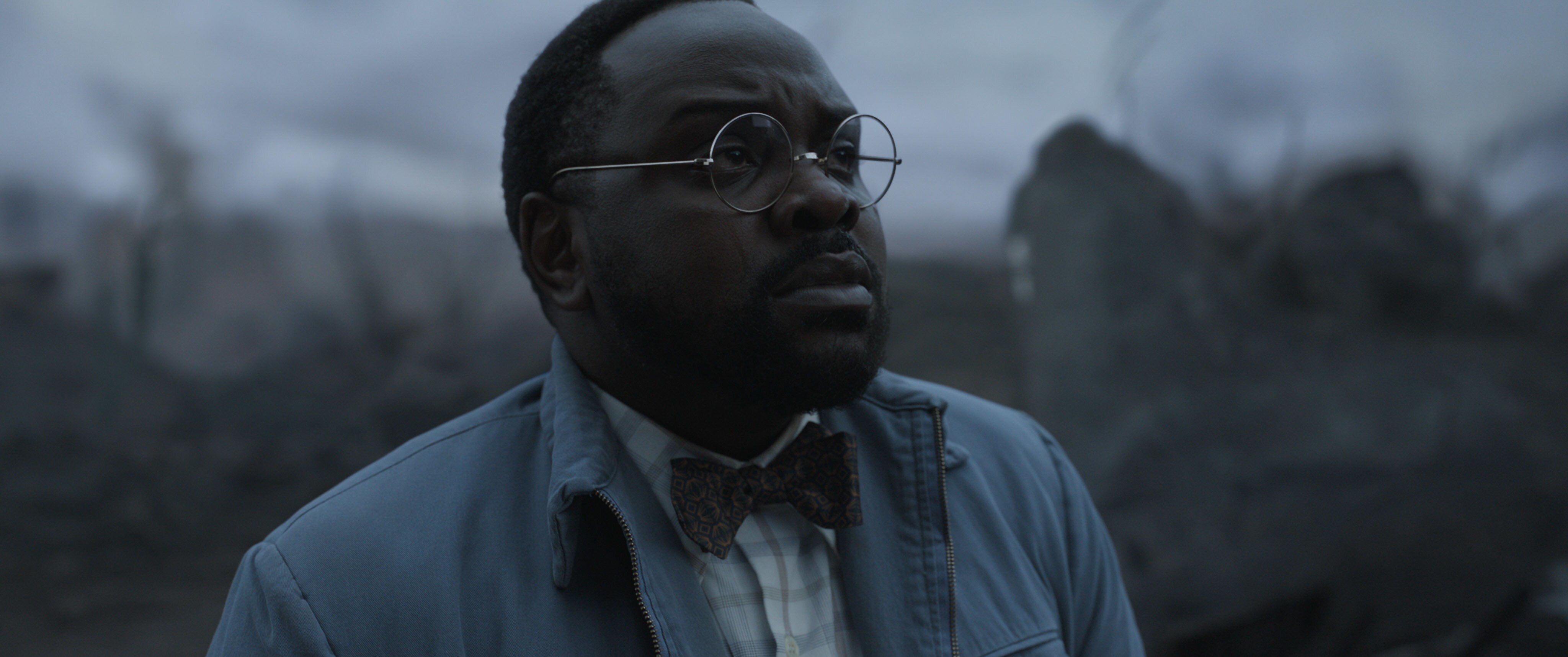 Closeup on Brian Tyree Henry as Phastos. He wears silver, circular wire-rim glasses, a light blue windbreaker, a plaid shirt, and a bow-tie.