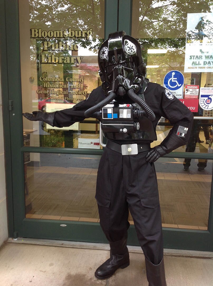 A cosplayer in a TIE fighter pilot costume stands in front of a library.