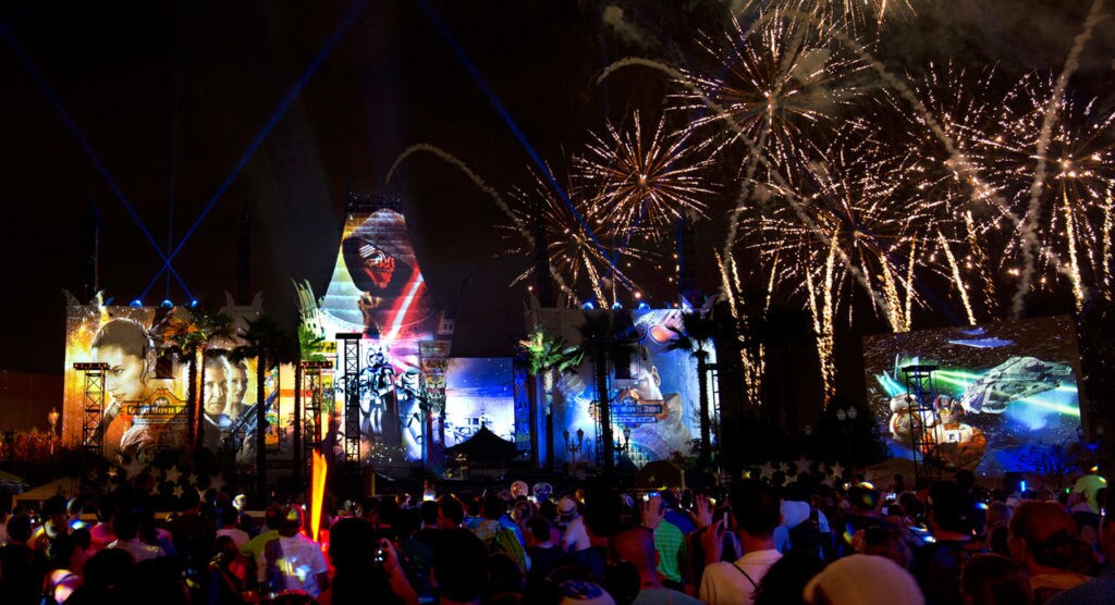 An enormous crowd of Star Wars fans at Galactic nights. A giant projector screen shows images from a Star Wars film, and fireworks light the sky.