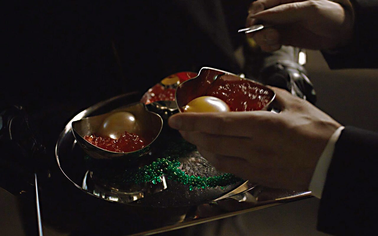 One hand holds a bowl of colo claw fish, which looks like a century egg next to caviar, and the other hand holds a fork in Solo: A Star Wars Story.