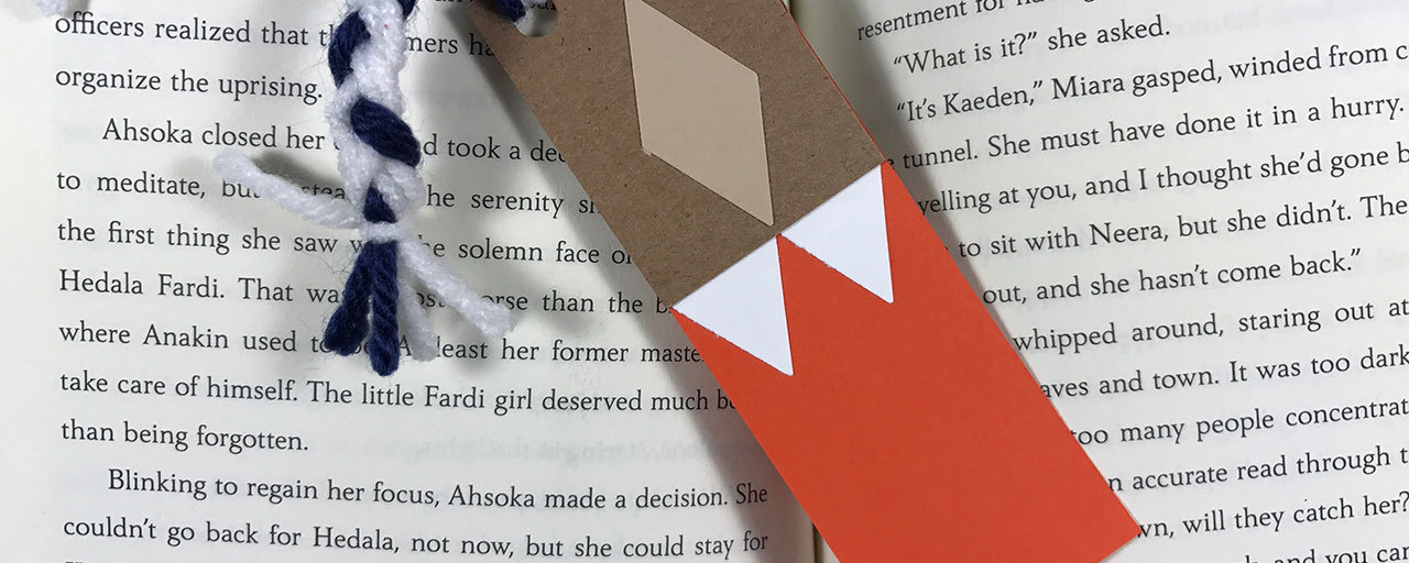 An Ahsoka Tano-inspired cardboard bookmark with yarn tassels, placed between the pages of a book.