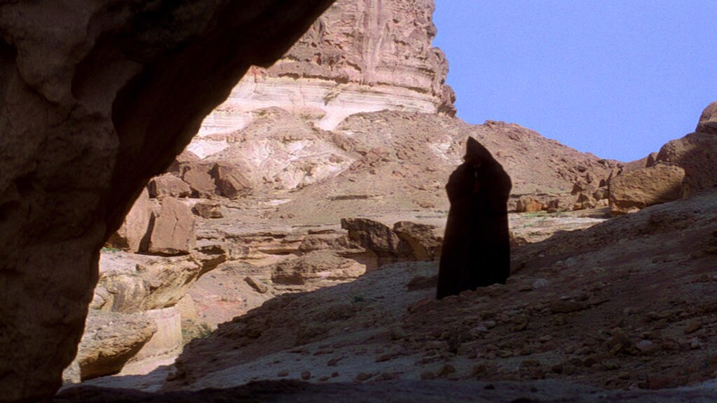 A cloaked Obi-Wan Kenobi walks into a canyon on Tatooine.