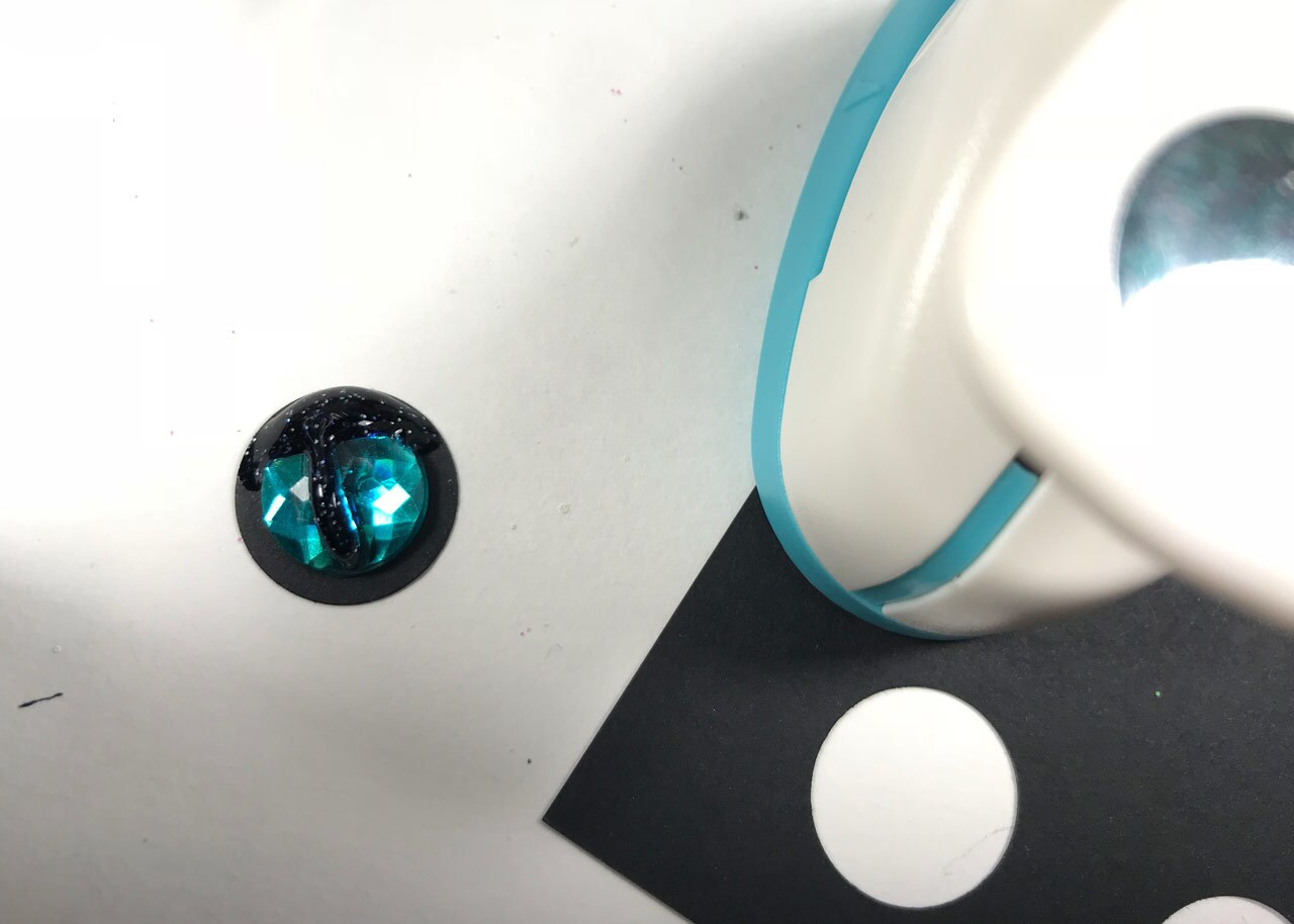 A blue gemstone next to hole-punched black cardstock. The gemstone will be used as an eye on a homemade, vulptex Christmas ornament.
