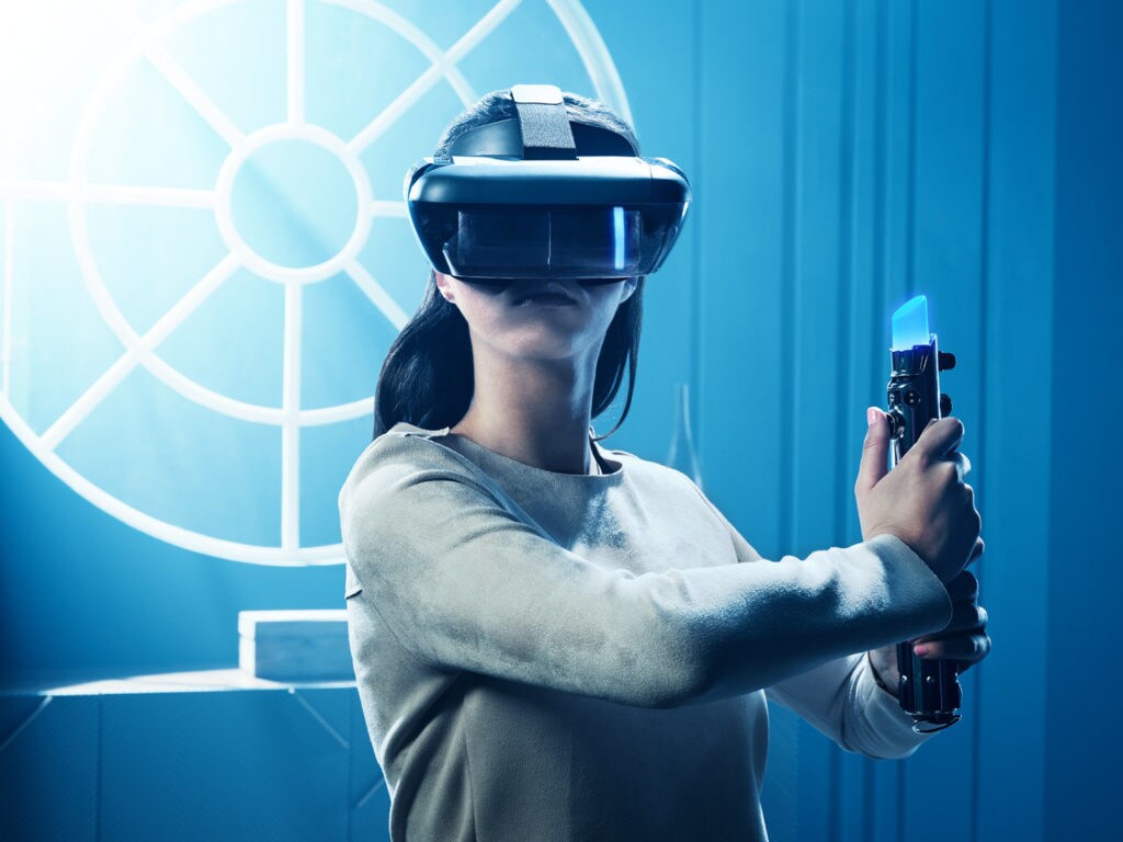 A woman wears a VR headset while wielding a lightsaber game controller for the Star Wars: Jedi Challenges augmented reality experience.