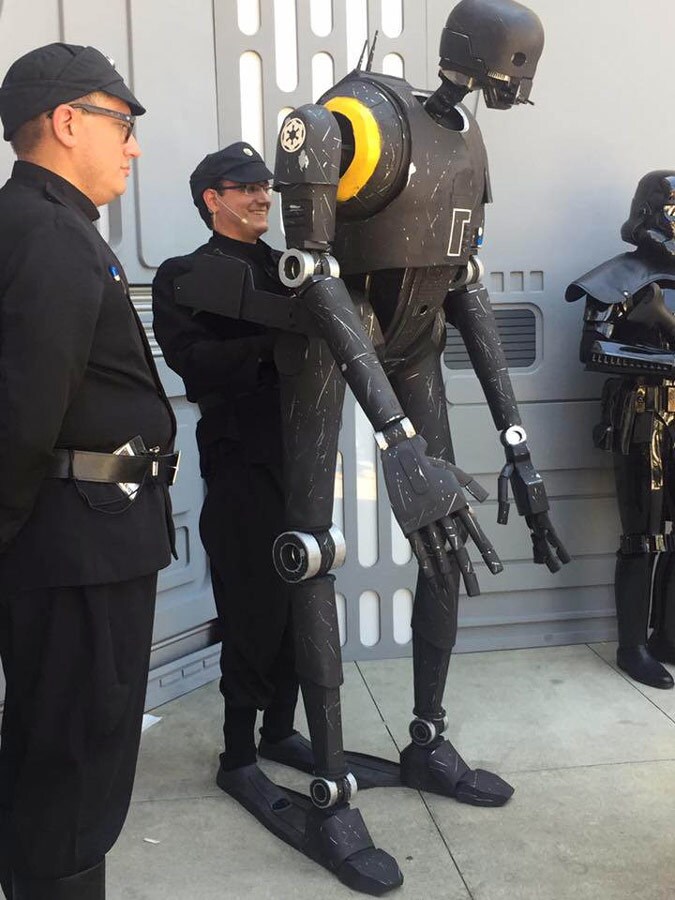 A Star Wars cosplayer, dressed in a black Imperial officer costume, holds up the life-size K-2SO puppet he created while fellow cosplayers stand beside him.