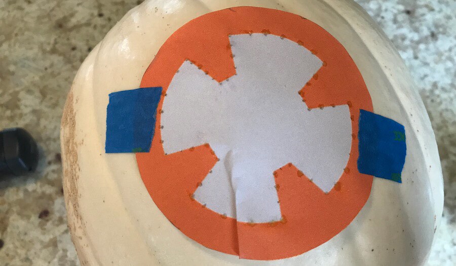 An orange, circular BB-8 design template taped onto a white pumpkin with carving guidelines.
