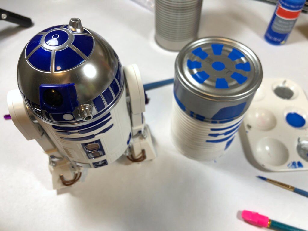 A toy R2-D2 next to a tin can painted to resemble him and eventually be part of a group of DIY wind chimes.