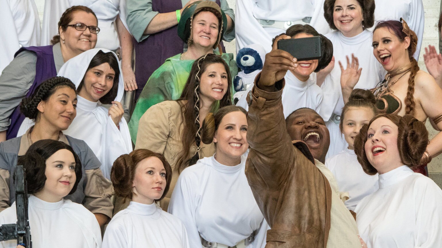 A Finn cosplayer takes a selfie with a large group of Princess Leia cosplayers.