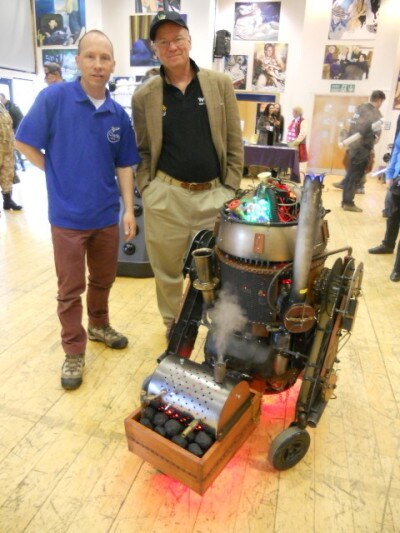 John Morton, the actor who played Dak Ralter in Star Wars: The Empire Strikes Back, poses with prop maker Mark Enright at a UK convention. They stand next to a steampunk inspired astromech droid that appears to be powered by coal.