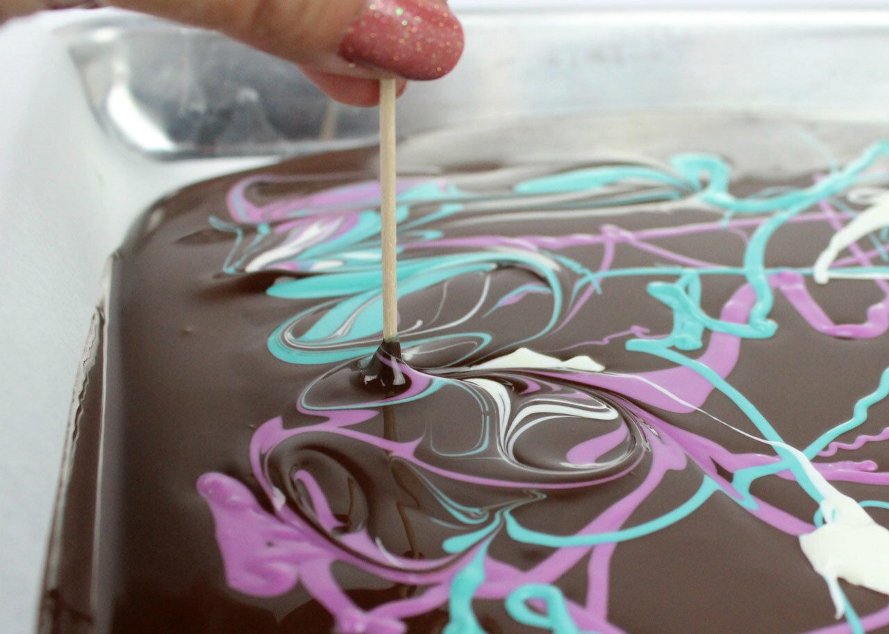 Spoonfuls of melted turquoise, lavender and white candy melts swirl into chocolate with a toothpick.