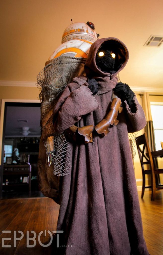 A cosplayer dressed as a Jawa carries BB-8 in a sack on their back.