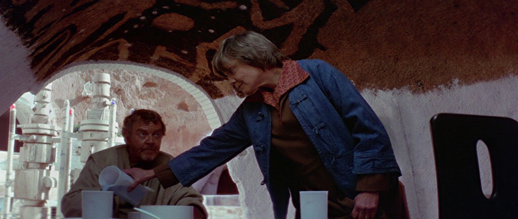 Beru Lars pours blue milk into a cup on the table as her husband, Owen, sits and eats in their home on Tatooine.