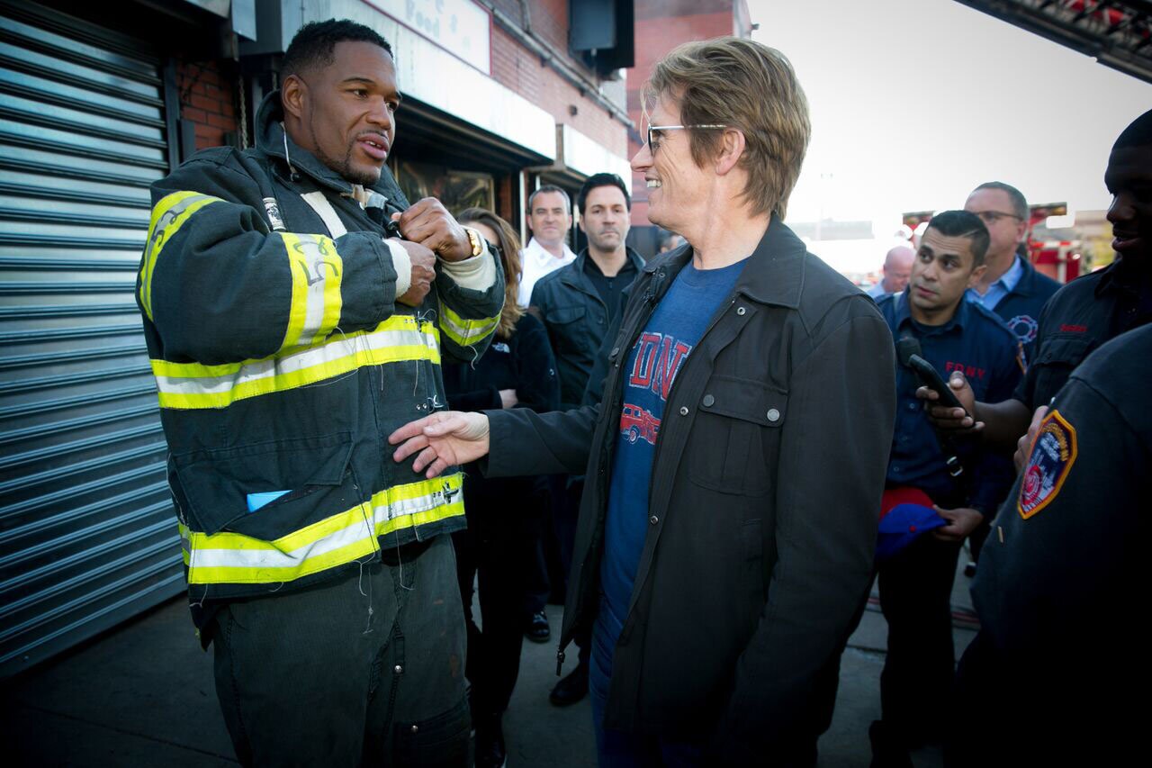 Denis Leary speaks with Michael Strahan, who is dressed as a firefighter, while members of the NYFD stand around them watching.
