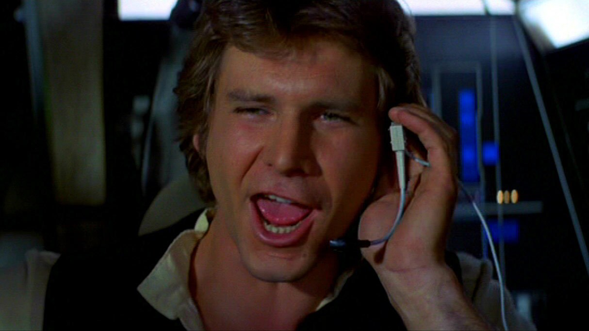 Han Solo smiles as he speaks into a headset while in the Millennium Falcon cockpit.