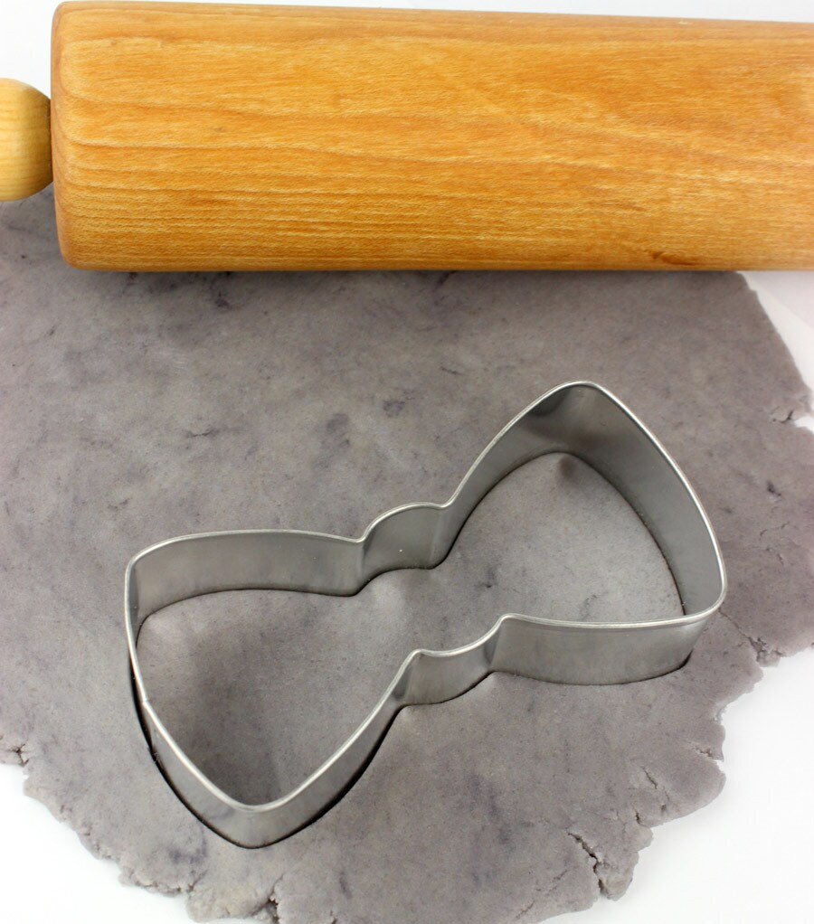 A bowtie cookie cutter pressed into cookie dough with a rolling pin above it.