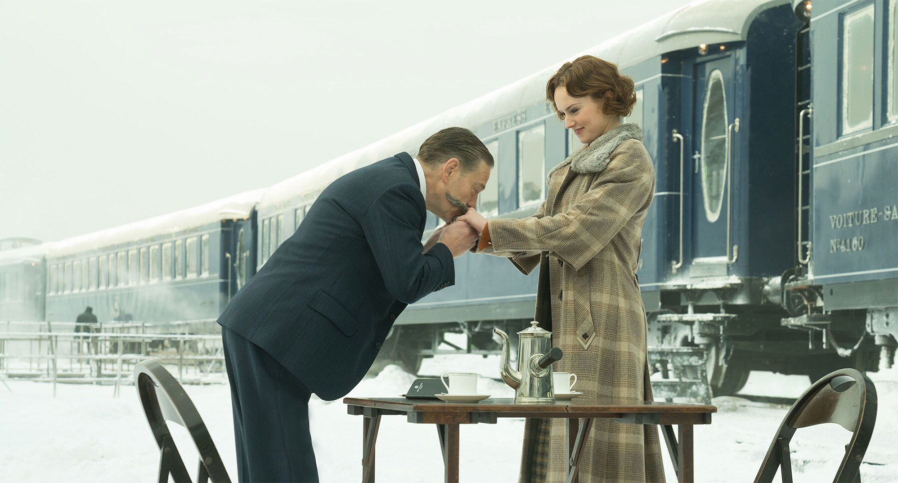 Kenneth Branagh (as Hercule Poirot) kissing  Daisy Ridley (as Mary Debenham) hands at a table set in "Murder on the Orient Express"