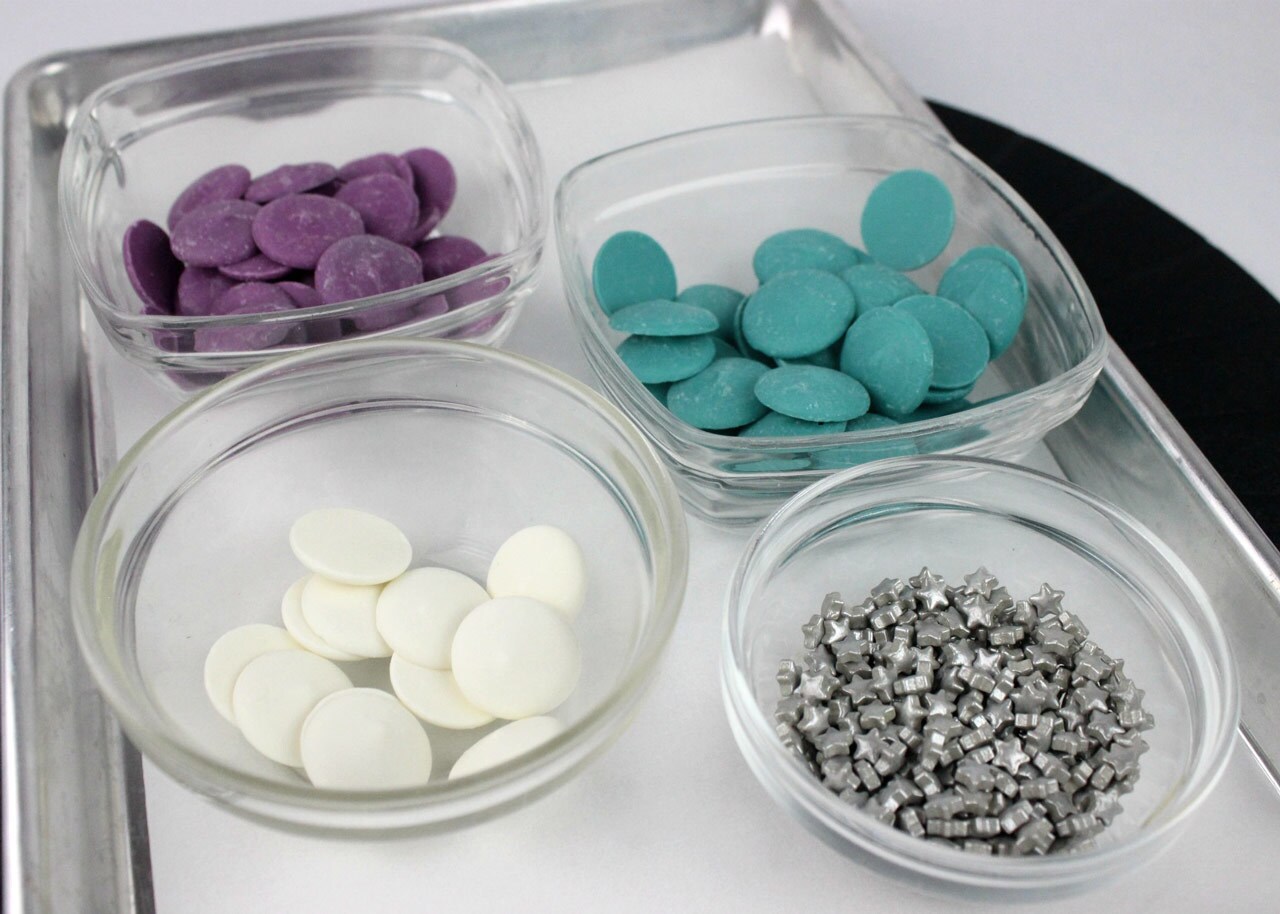 A trey of turquoise, lavender and white candy melts in separate bowls beside a bowl of silver stars.