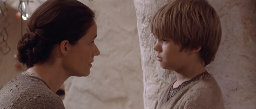 Shmi Skywalker looks at young Anakin Skywalker in their home.